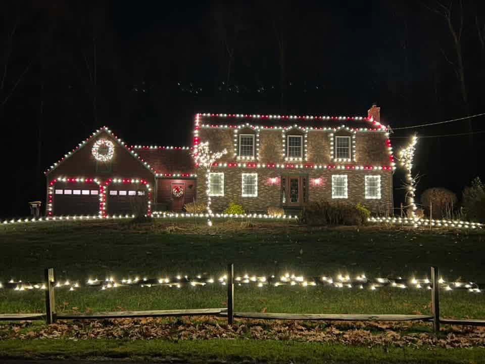 Brick two-story home with red and warm white Christmas lights, a lit wreath, and a fence lined with lights