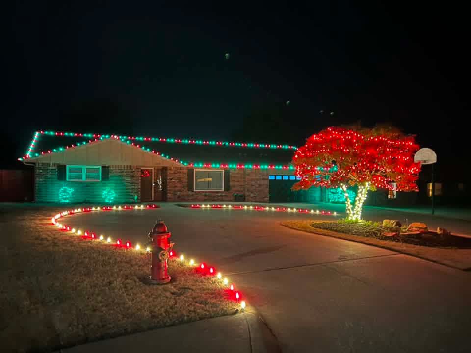 Ranch home with red and green roofline lights, a red-lit tree, and driveway lined with lights